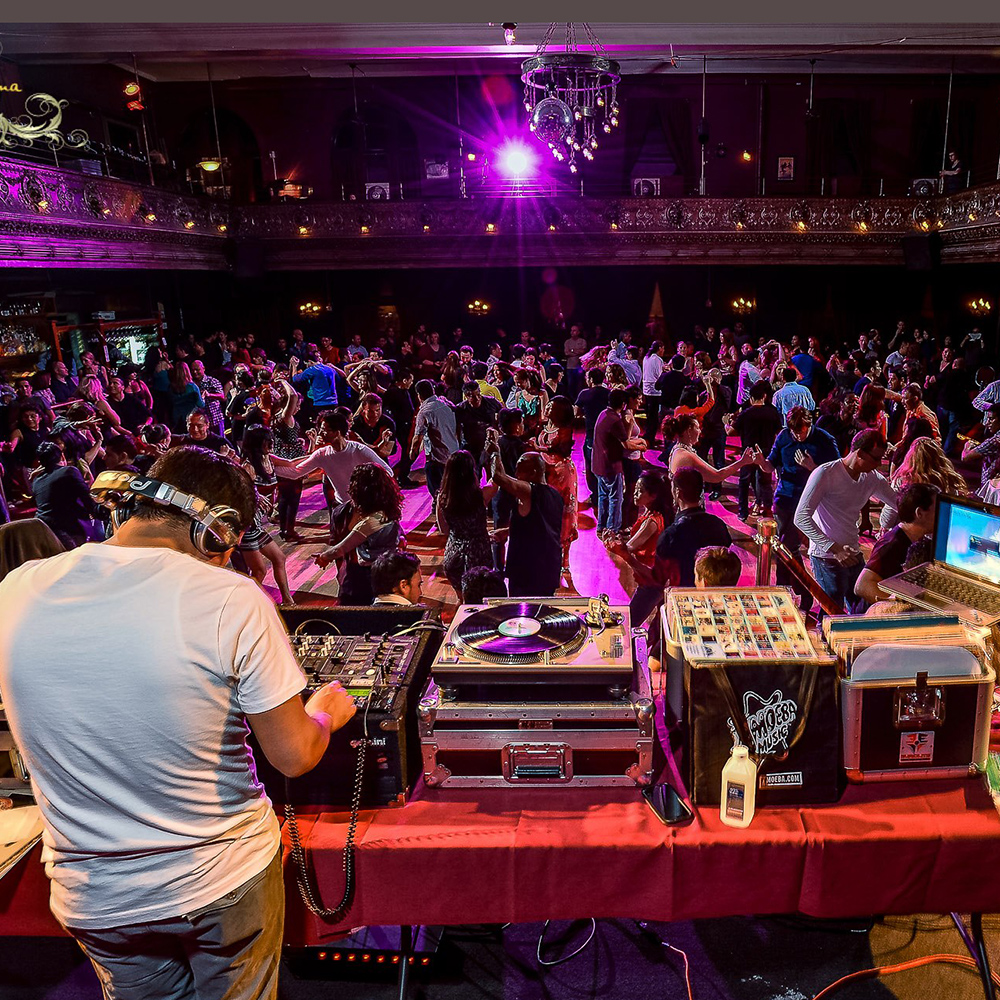 A DJ plays vinyl salsa music from the stage while dancing couples pack the dance floor