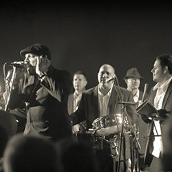 A black-and-white photo of a man singing into. microphone while a band plays instruments behind him